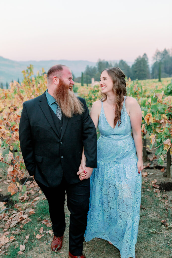 couple smiling at each other holding hands in vineyard for their engagement session in napa valley california at teh castello di amorosa by Sarah Schweyer Photography