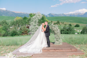 bride and groom kissing under floral arch on their wedding day with a mountain view behind them at Foster Creek Farms in Bozeman Montana
